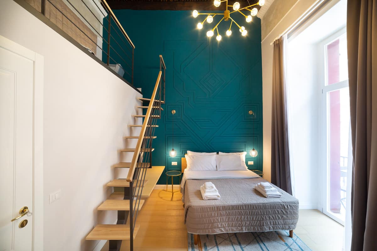 Une chambre à coucher avec un lit, un escalier, un lustre et une fenêtre avec des rideaux.