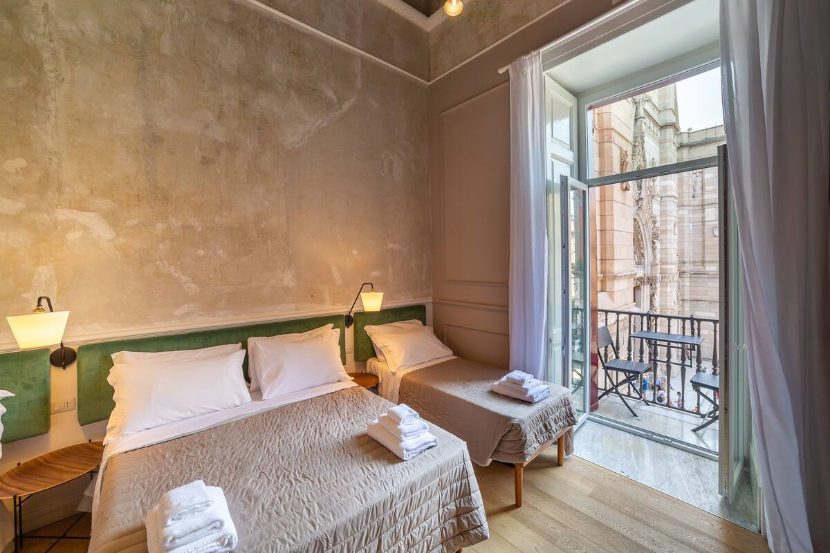 Une chambre d’hôtel avec deux lits, un balcon donnant sur un immeuble, ainsi qu’une petite table et une chaise placées à l’extérieur.