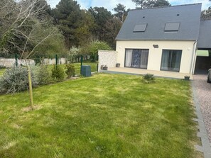 Une maison avec un toit en pente, un petit jardin devant avec de la pelouse, un mur de pierres et une zone clôturée avec des arbres et des arbustes.