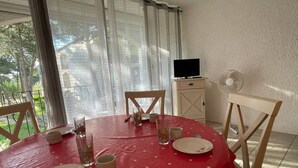 A dining area with a round table covered in a red tablecloth with white polka dots, surrounded by wooden chairs. There are plates, cups, and cutlery on the table. A television is mounted on the wall to the right, and a fan is on the floor to the right of the table. - **Cozy Seaside Studio with Balcony, 200m from Beach, Sleeps 4** (Argelès-sur-Mer)