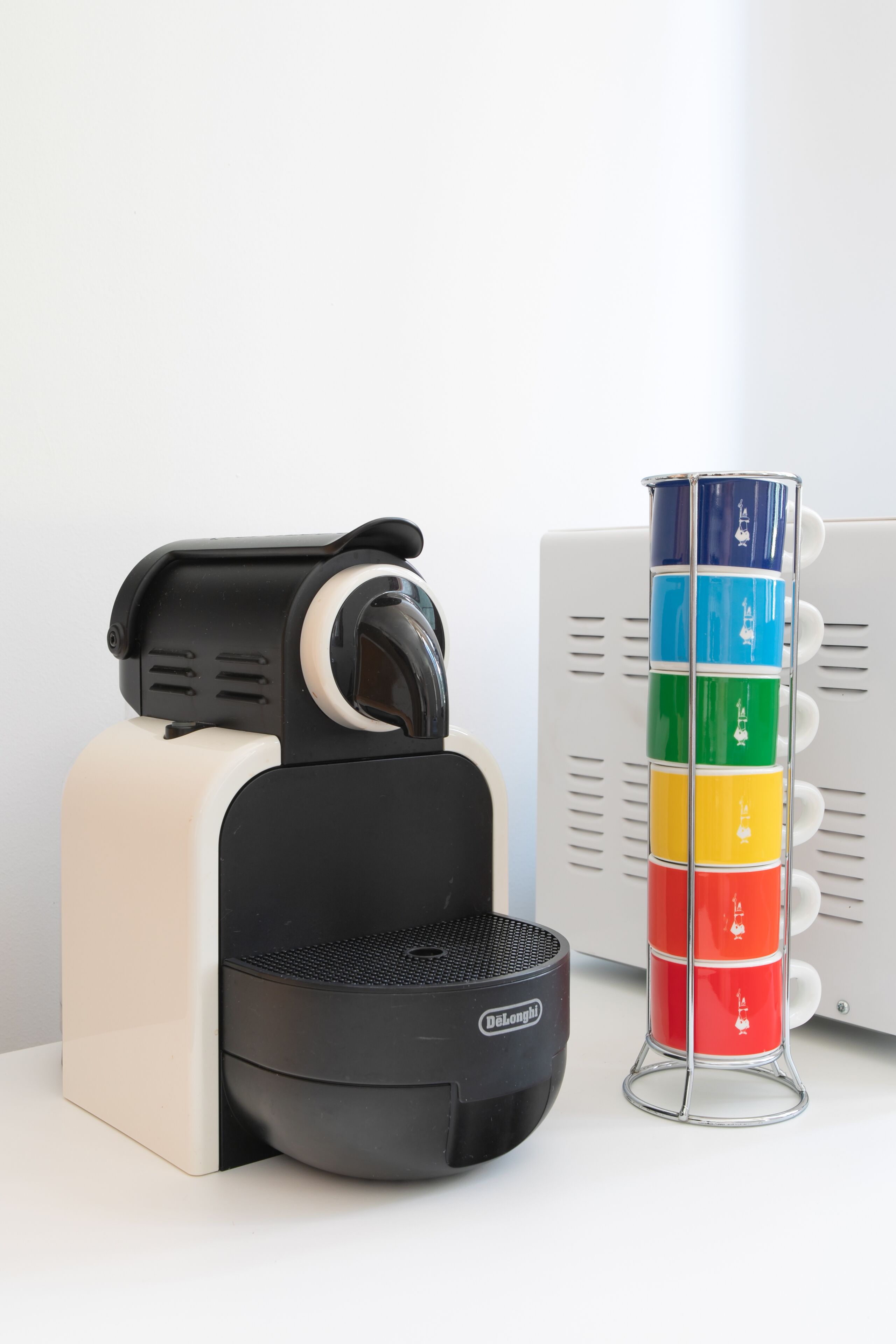 A coffee maker with a black and white design, a stack of colorful cups, and a white wall in the background.