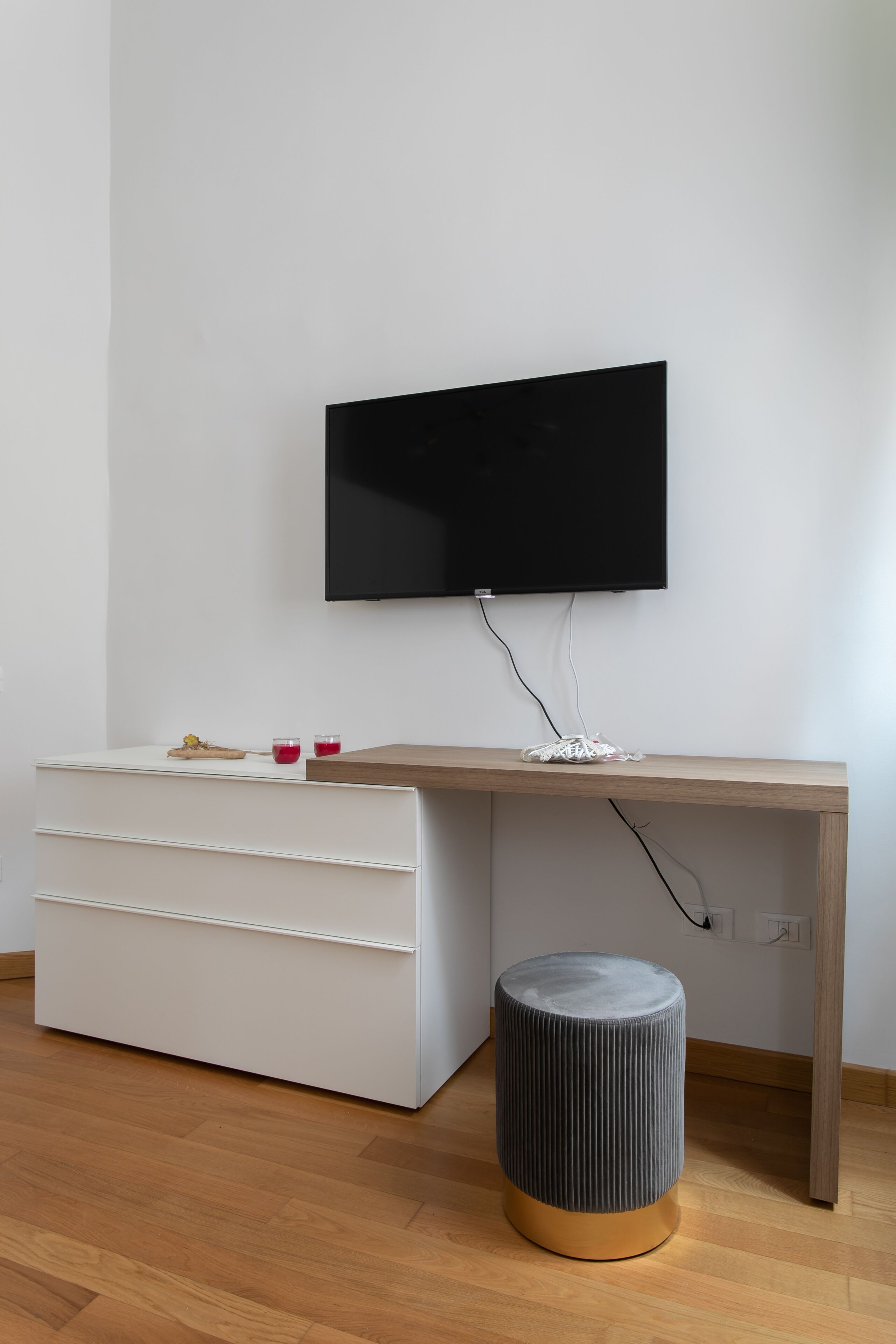 Una camera moderna con una scrivania in legno, una TV a schermo piatto montata a parete, un mobile contenitore bianco e un poggiapiedi grigio.