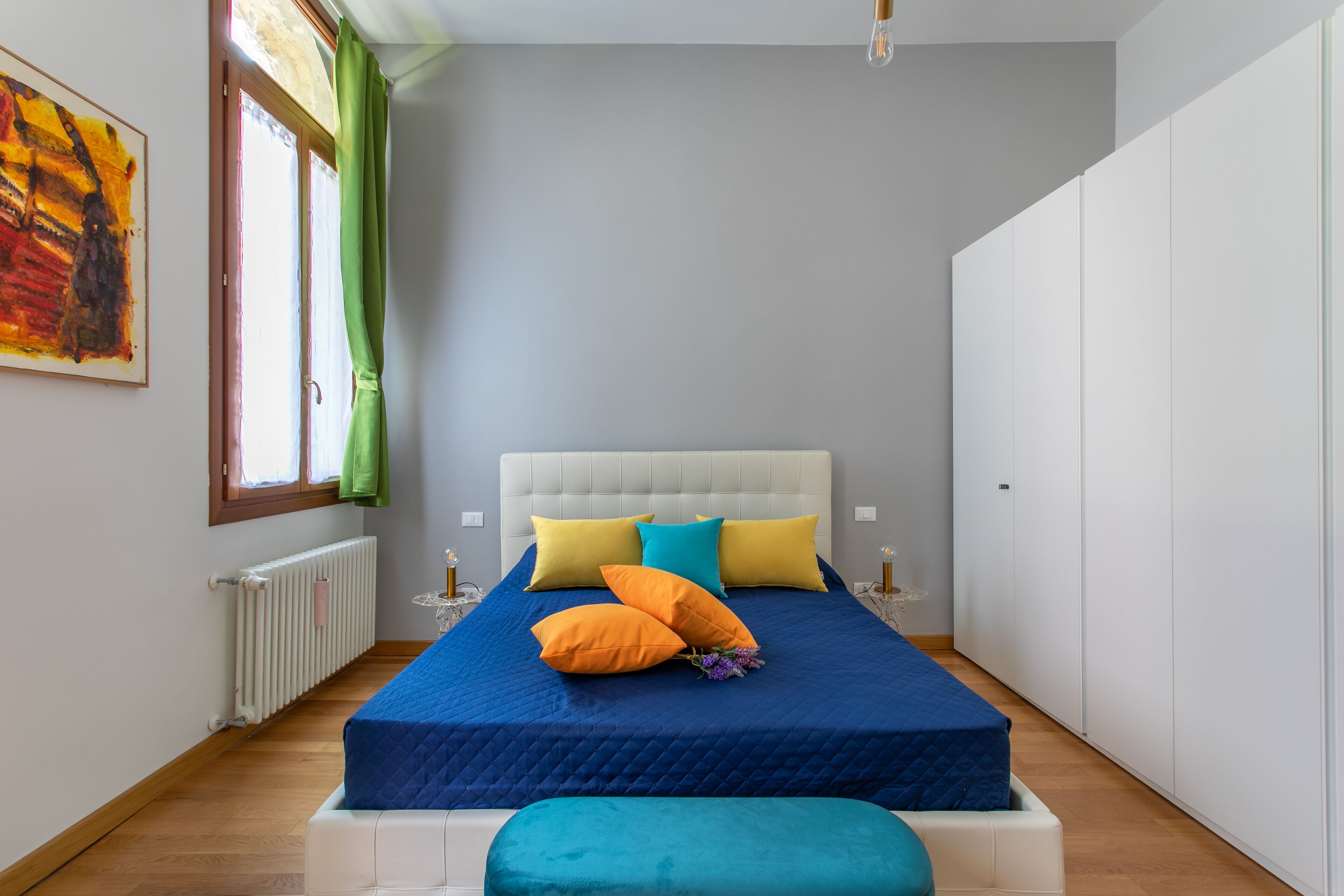 Un dormitorio con cama, almohadas, un radiador, una ventana con cortinas y un armario.