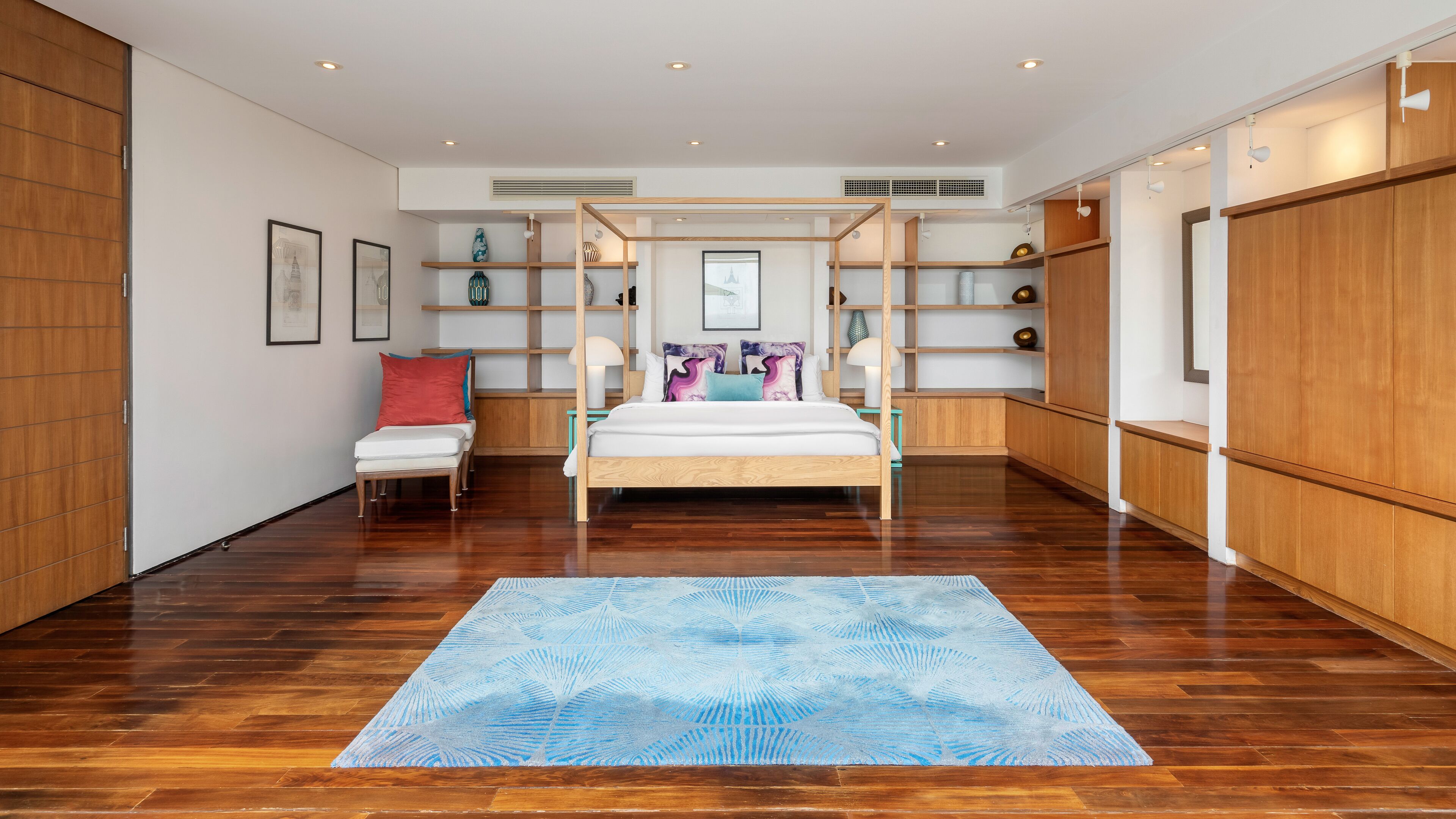 A modern bedroom with a wooden four-poster bed, a small seating area with a red pillow, and built-in shelving with decorative items.