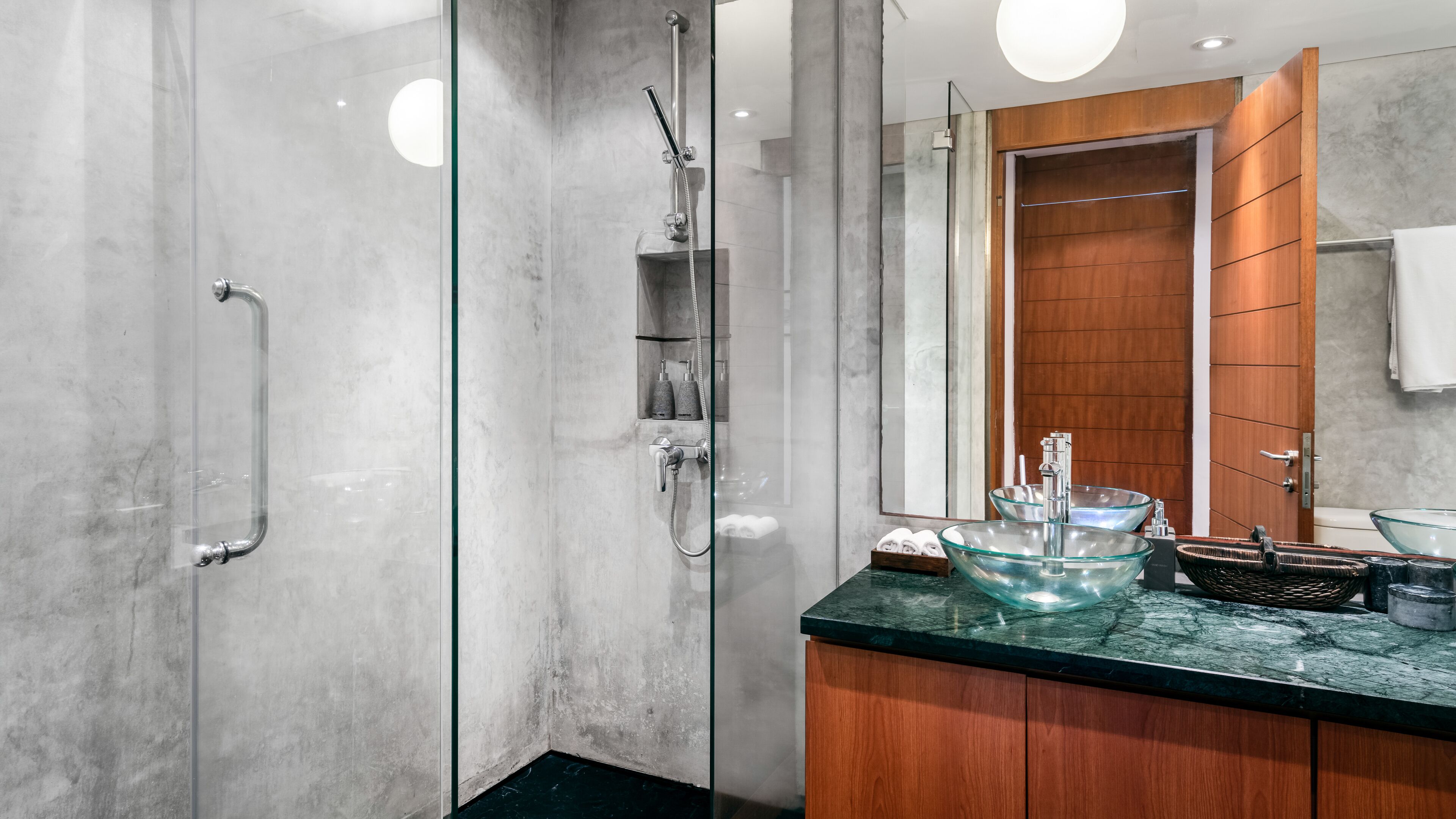 Un baño moderno con ducha de vidrio, tocador de madera y lavabo de vidrio, además de un espejo grande.