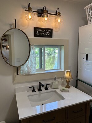 Un baño con un lavabo blanco, grifos negros, un espejo redondo y una ventana con vistas a la vegetación. Hay un letrero sobre el espejo que dice “BEGIN EACH DAY with a Smile.”