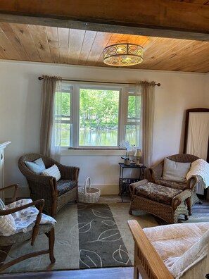 A cozy room with wicker furniture, a large window with curtains, a wooden ceiling, and a table with a lamp.