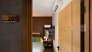 A narrow hallway leading to a bedroom with a brick wall, a bed with green pillows, a desk with a lamp, and a wardrobe. - Cove W Suites (Jakarta)