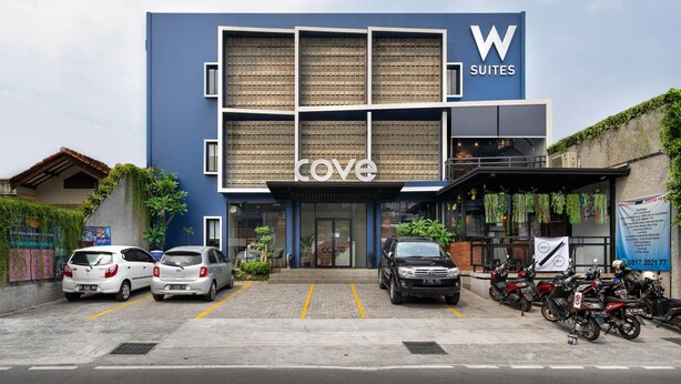 A modern building with a blue facade, a 'W Suites' sign, and a 'Cove' sign. There are cars and motorcycles parked in front. - Cove W Suites (Jakarta)
