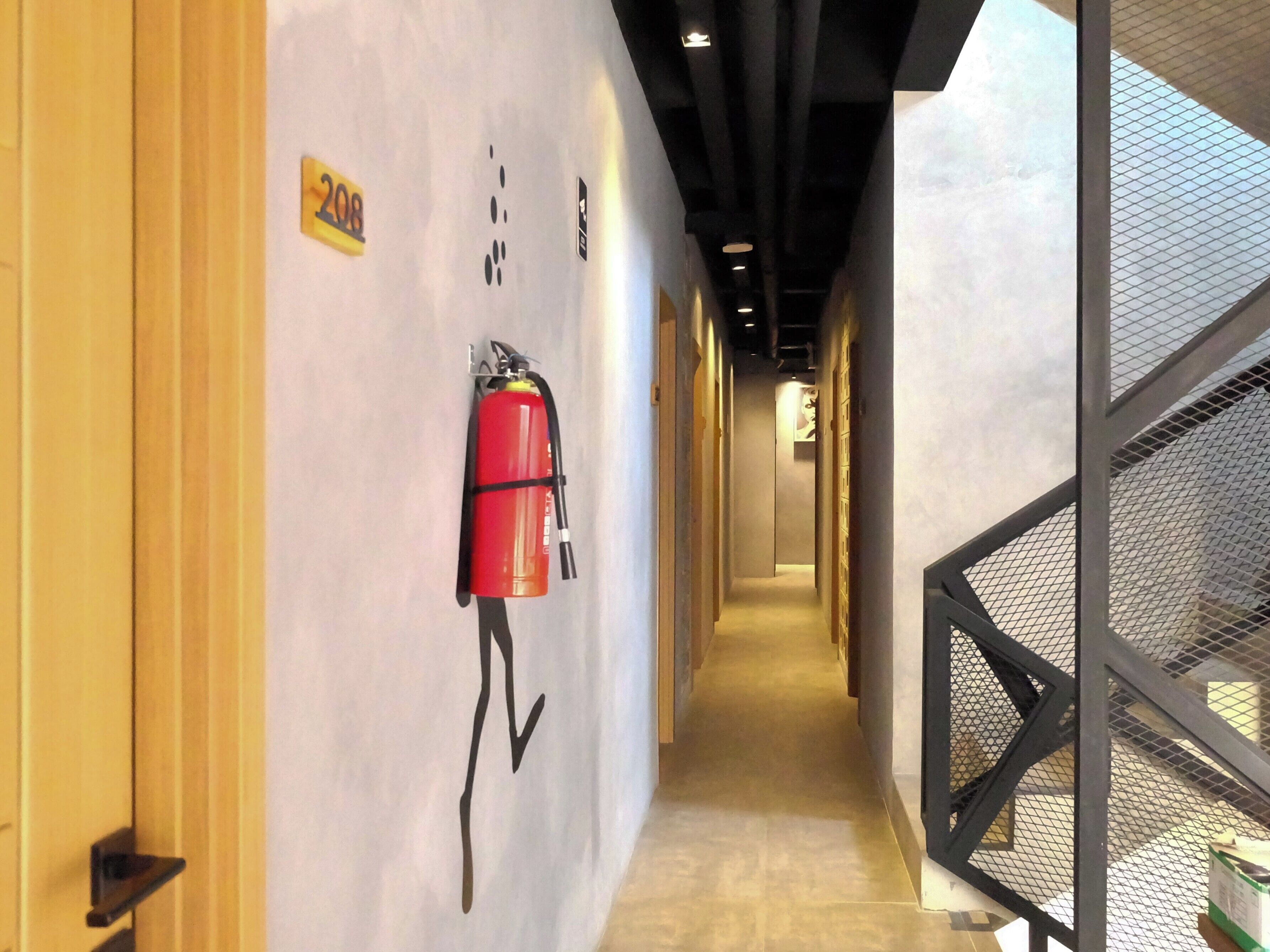 A hallway with a fire extinguisher mounted on the wall, a number '208' sign, and a mesh railing on the right.