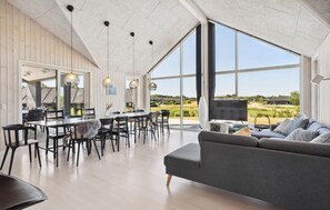 Un vaste espace de vie avec une grande fenêtre, un coin repas comprenant des chaises et une table, et un canapé gris.