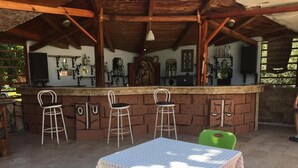 Un espace bar extérieur doté d’un plafond en bois rustique, d’un comptoir avec des tabourets et d’une table recouverte d’une nappe à carreaux.