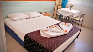 Un lit bien fait, avec du linge de lit blanc et une couvre-lit marron à motifs. On y trouve une petite table recouverte d’une nappe blanche et un vase rempli de fleurs.