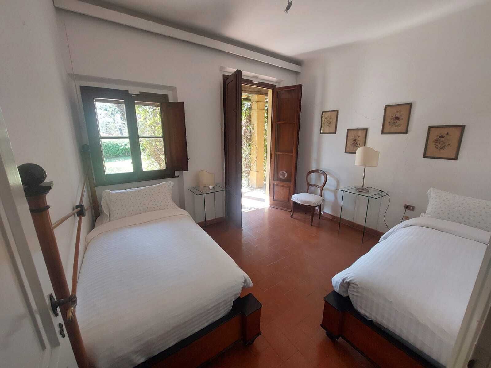 A bedroom with two beds, a chair, a small table with a lamp, and a door leading to an outdoor area.