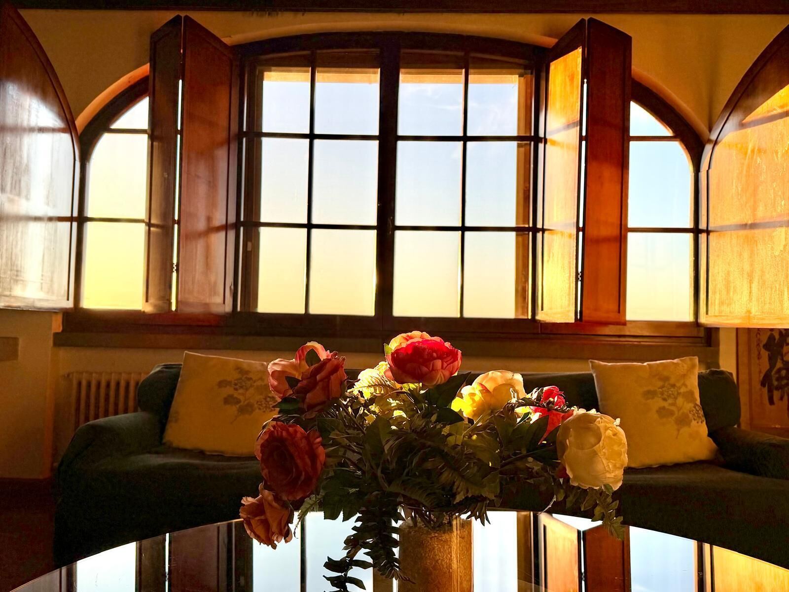 Een kamer met een ronde glazen tafel, een groene bank en een raam met houten luiken.