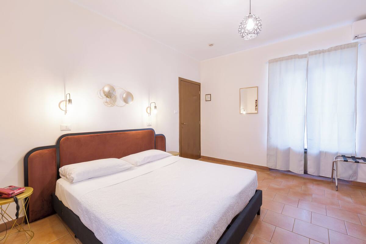 A neatly made bed in a hotel room with a wooden headboard, a bedside table with a lamp, and a window with curtains.