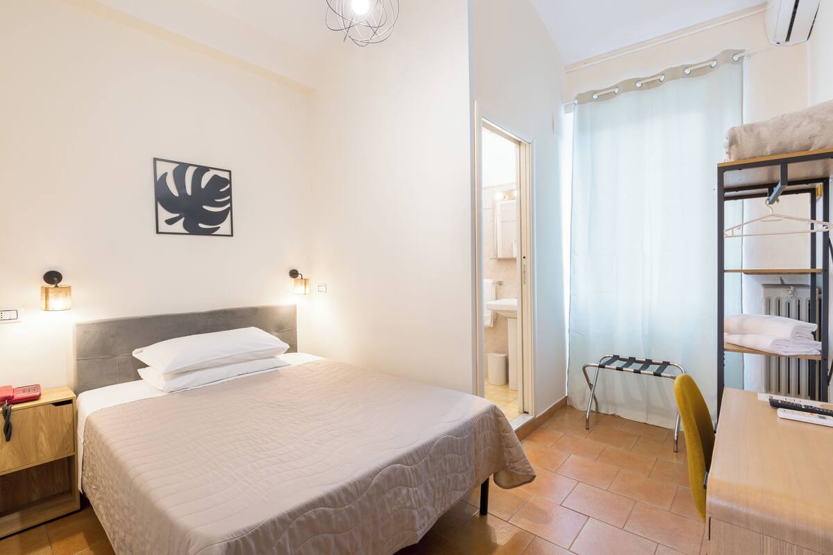 A hotel room with a bed, bedside table, lamp, and a view of the bathroom.
