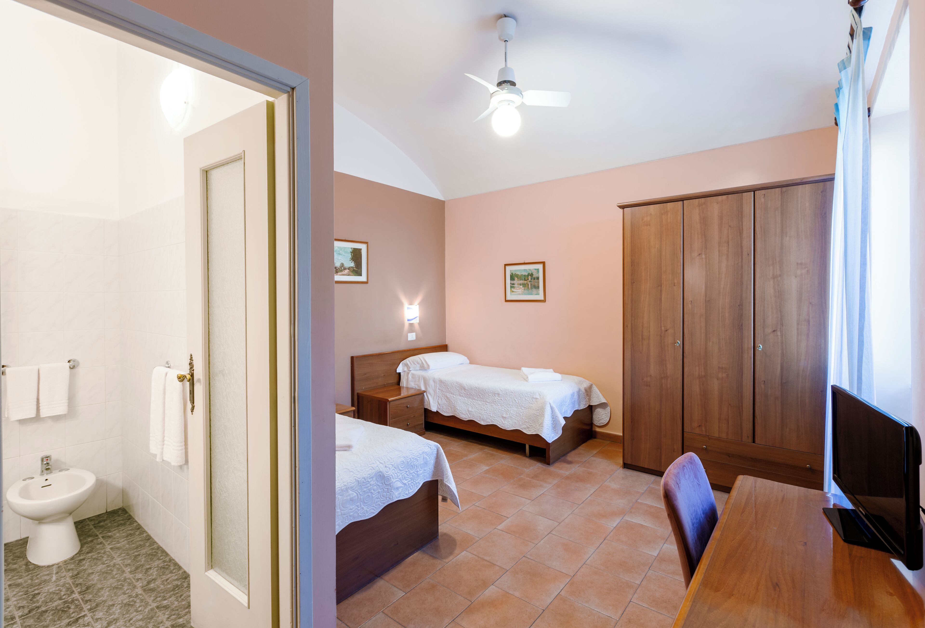 A bedroom with two beds, a wooden wardrobe, a television, and a bathroom with a toilet and bidet.