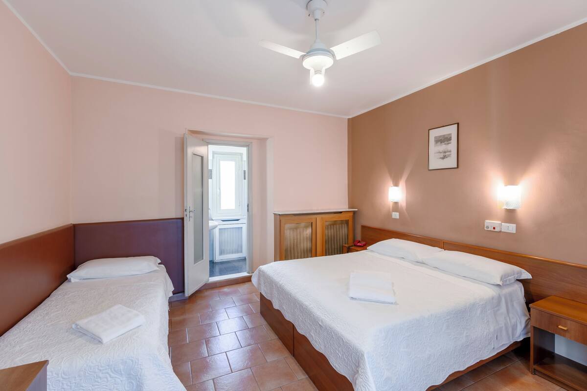 A hotel room with two beds, a ceiling fan, a door leading to a balcony, and wall-mounted lights.