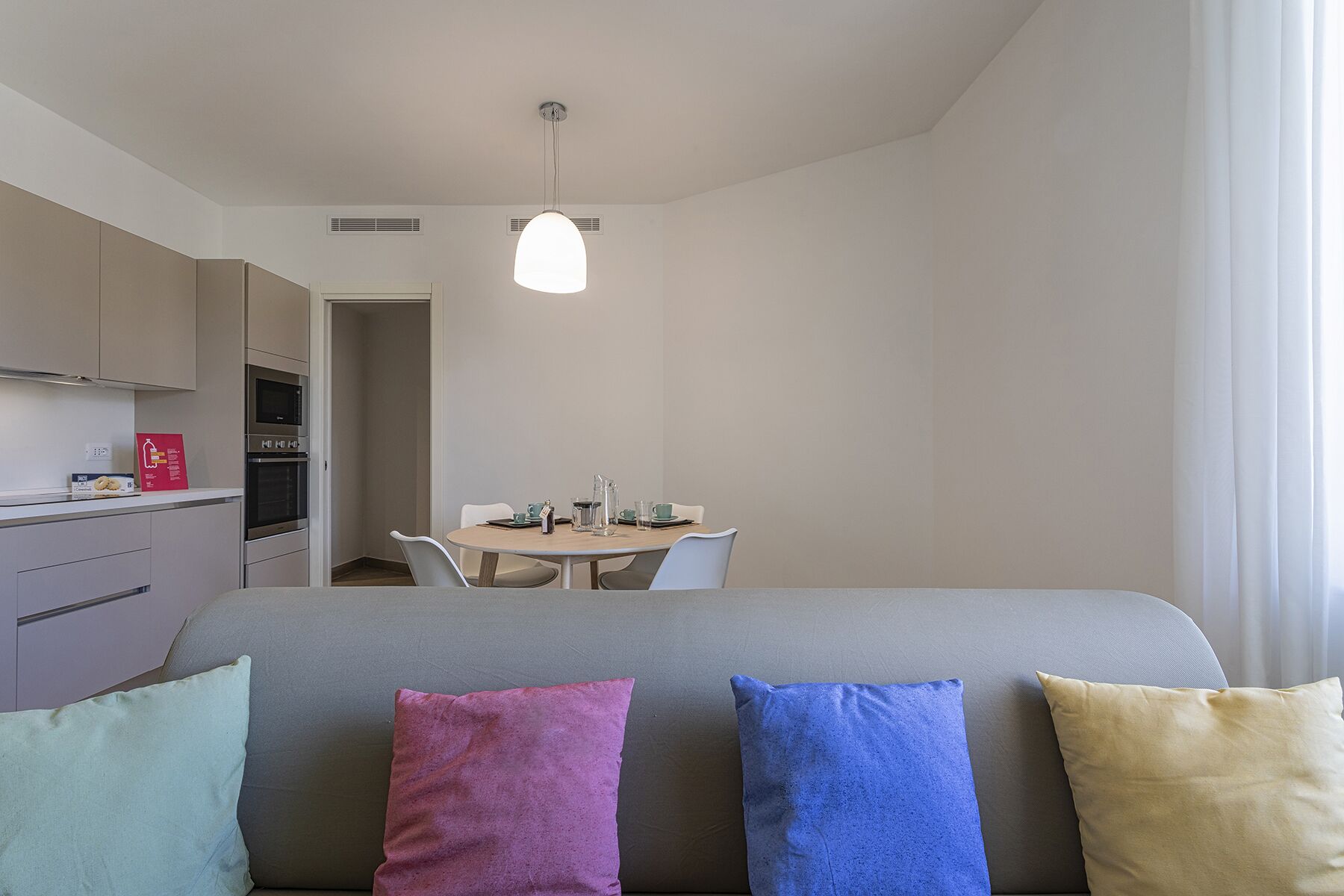 Una habitación de hotel moderna con un sofá gris, cojines coloridos, una mesa de comedor con sillas, una kitchenette y una lámpara colgante.