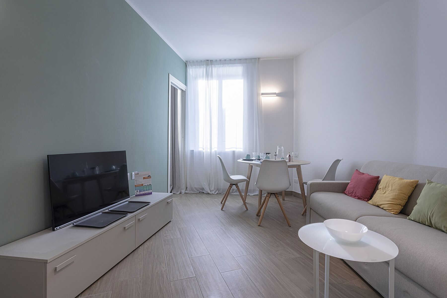 A modern living room with a flat-screen TV, a dining table with chairs, a sofa with cushions, and a small white table.