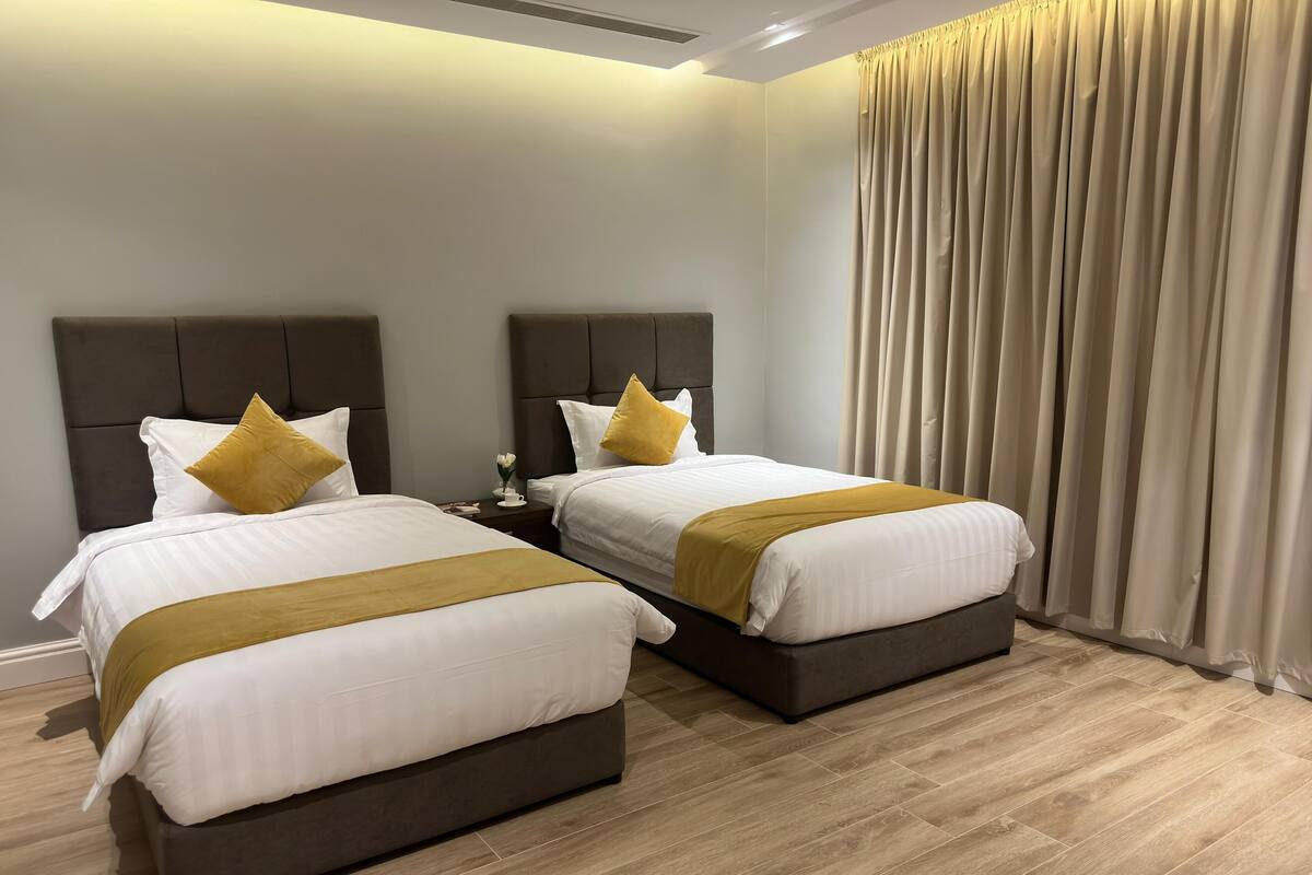 A hotel room with two beds, each with a headboard, white bedding, and a mustard-colored throw. The room has a wooden floor and a large window with beige curtains.
