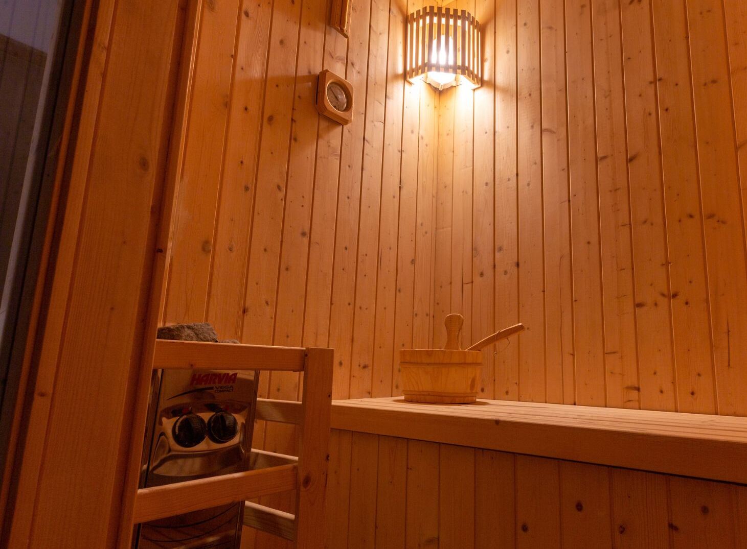 A wooden cabin interior with a wooden bench, a small table with a wooden cup and a spoon, and a Harvia brand bottle.
