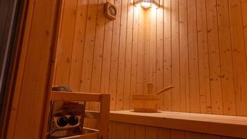 A wooden cabin interior with a wooden bench, a small table with a wooden cup and a spoon, and a Harvia brand bottle.