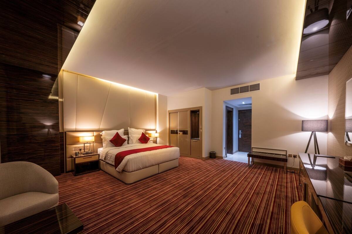 A modern hotel room with a large bed, bedside tables, a chair, and a desk with a lamp.