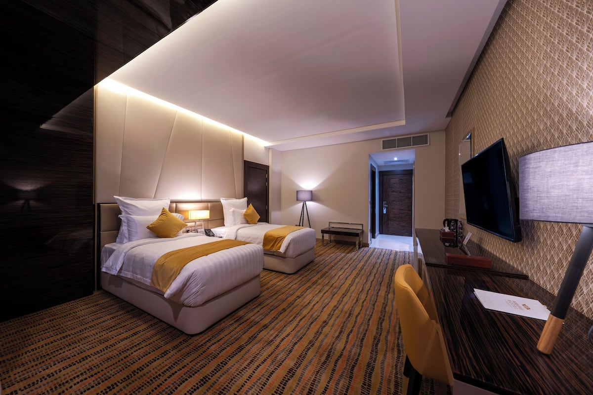 A hotel room with two beds, a desk, a chair, a TV, and a lamp.