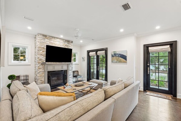 A modern living room with a stone fireplace, a flat-screen TV mounted above it, a large sectional sofa, and a dining area with a coffee table.
