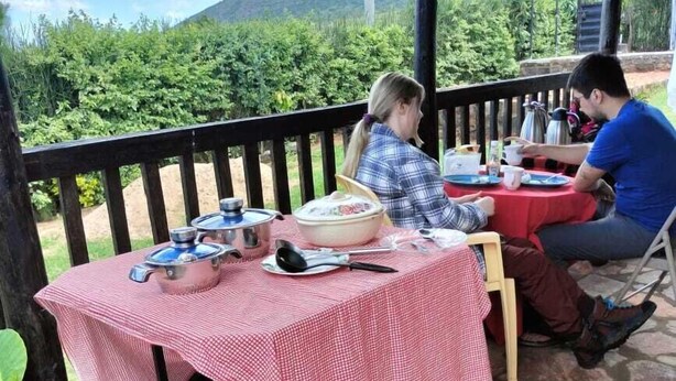 A covered outdoor dining area with a red checkered tablecloth, stone flooring, and a view of greenery and hills in the background.