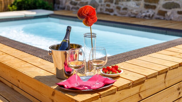 Une terrasse en bois avec une piscine, une bouteille de champagne, deux verres à vin, un saladier de fraises et une fleur rouge.