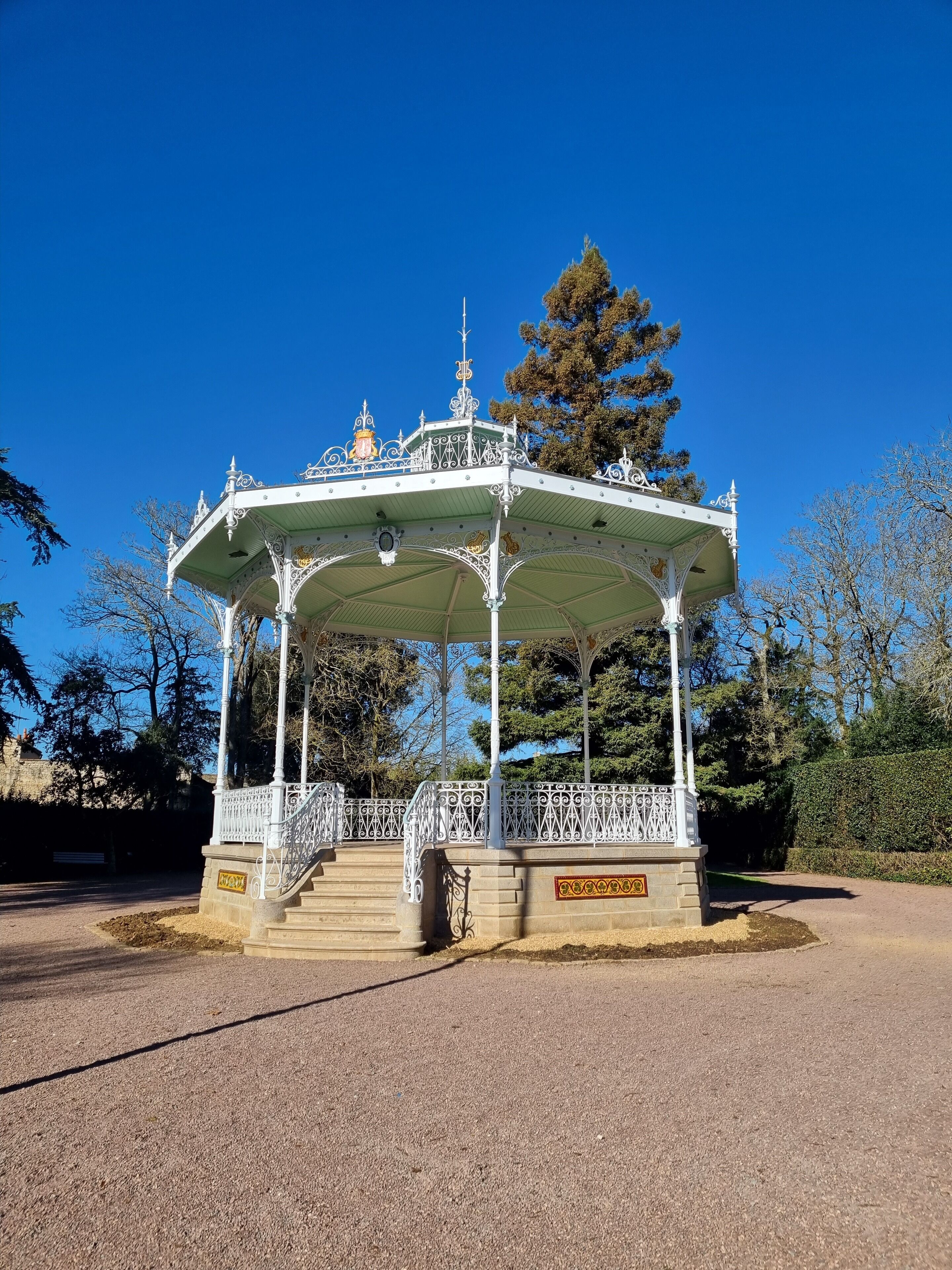 A white gazebo with ornate metal railings and a green roof, surrounded by trees and a clear blue sky.