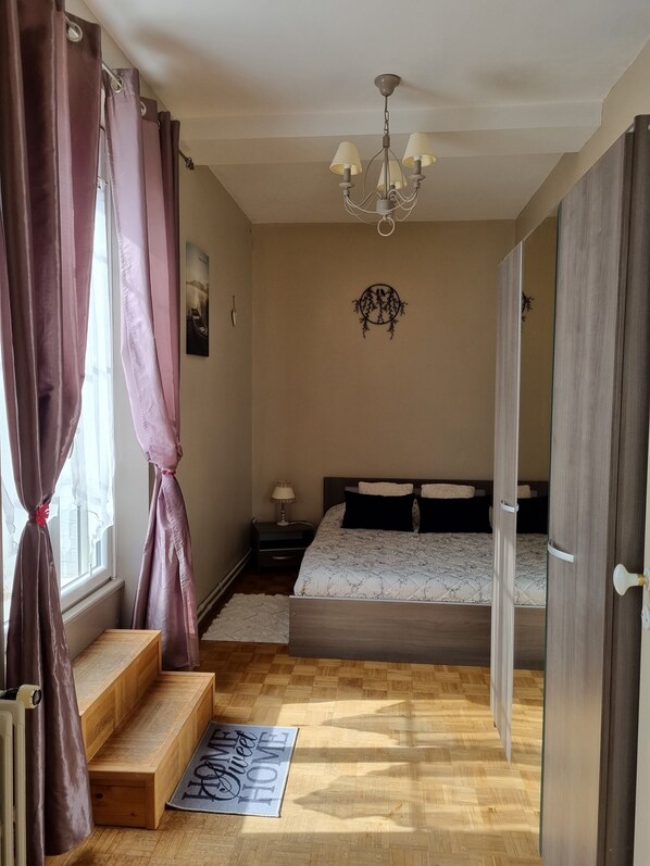 Un dormitorio con cama, mesita de noche, lámpara, candelabro, ventana con cortinas, puerta y un pequeño banco de madera.
