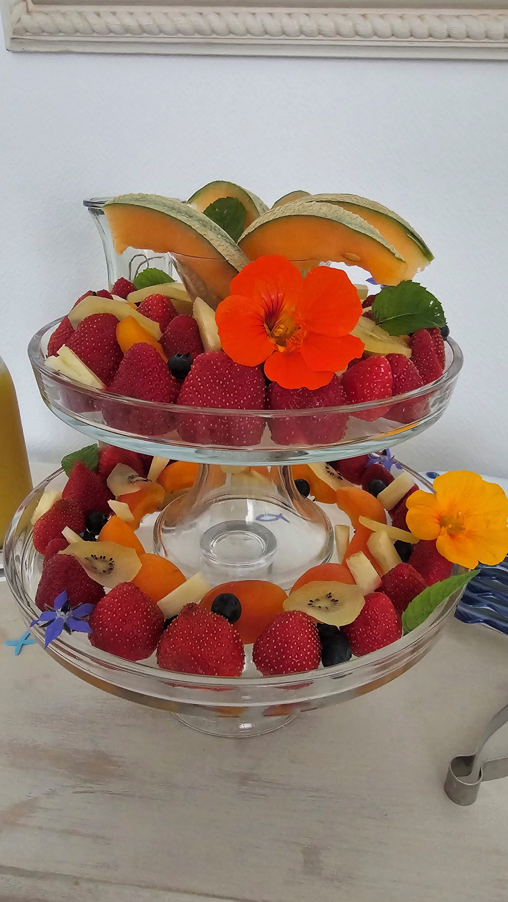 A two-tiered glass fruit stand with strawberries, blueberries, kiwi, and melon slices.