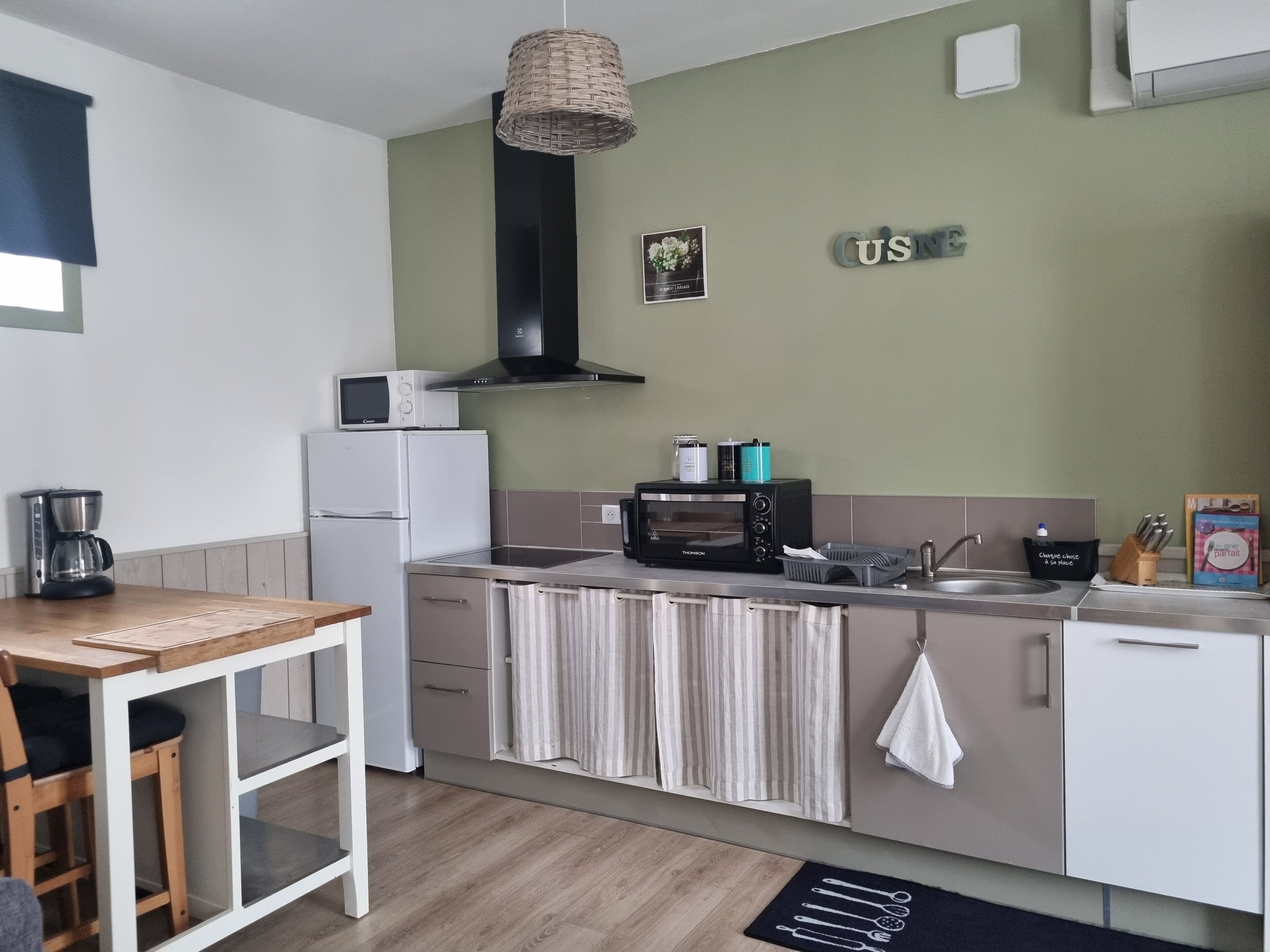 Une cuisine compacte équipée d’un four à micro-ondes, d’un grille-pain et d’une machine à café. On y trouve une table à manger avec des chaises, un luminaire suspendu et une serviette accrochée à la porte du four.