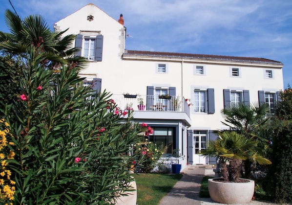 A large white building with multiple windows and shutters, surrounded by lush greenery and potted plants.