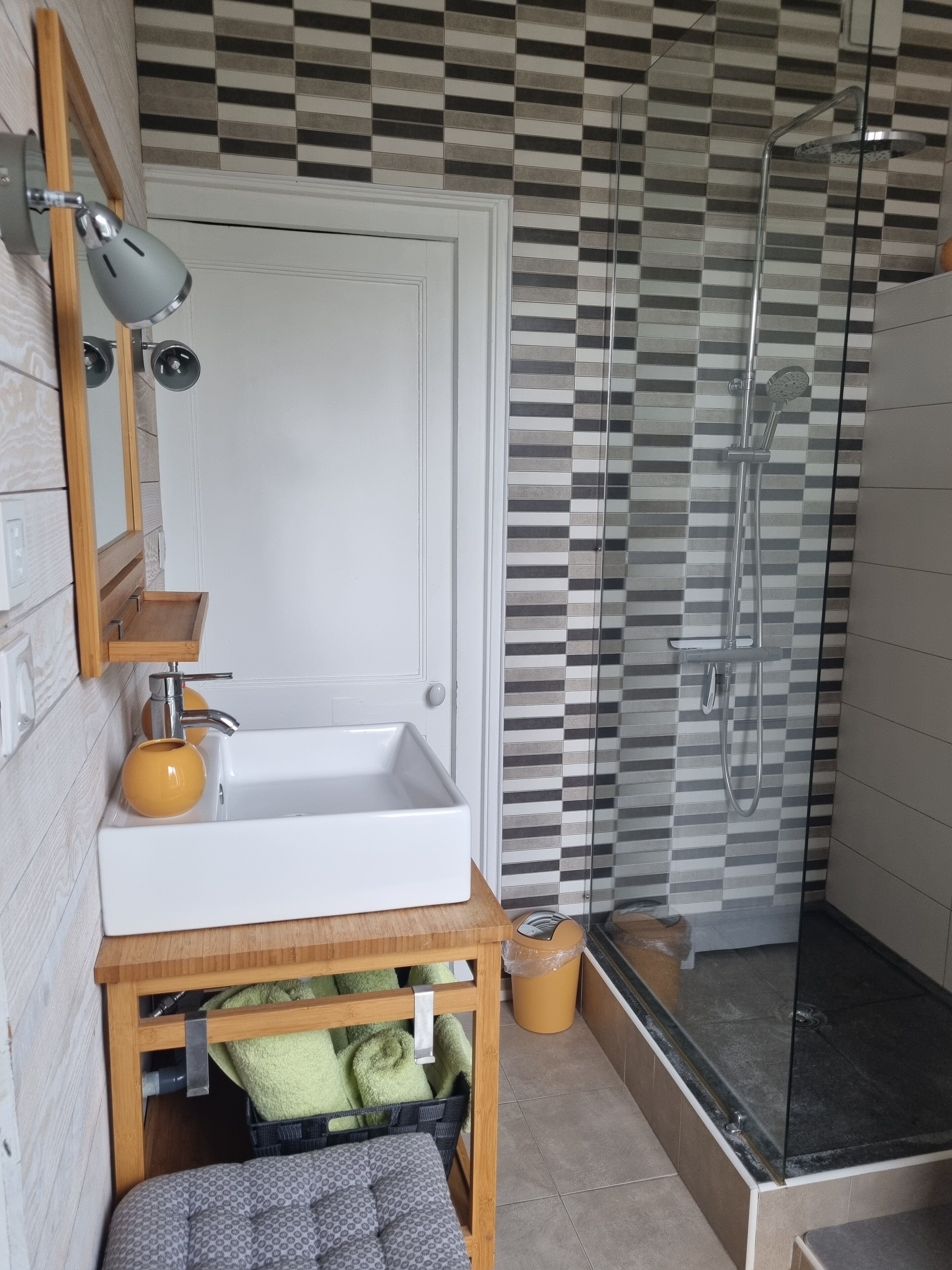 Une salle de bain avec un lavabo blanc, un miroir, un porte-serviettes et un espace douche dont le mur est recouvert de carreaux à motifs.