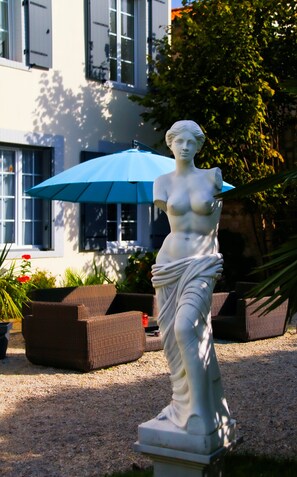 Una estatua de una mujer en un jardín, con un paraguas azul, muebles de mimbre y un edificio al fondo.