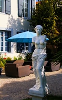 Una estatua de una mujer en un jardín, con un paraguas azul, muebles de mimbre y un edificio al fondo.