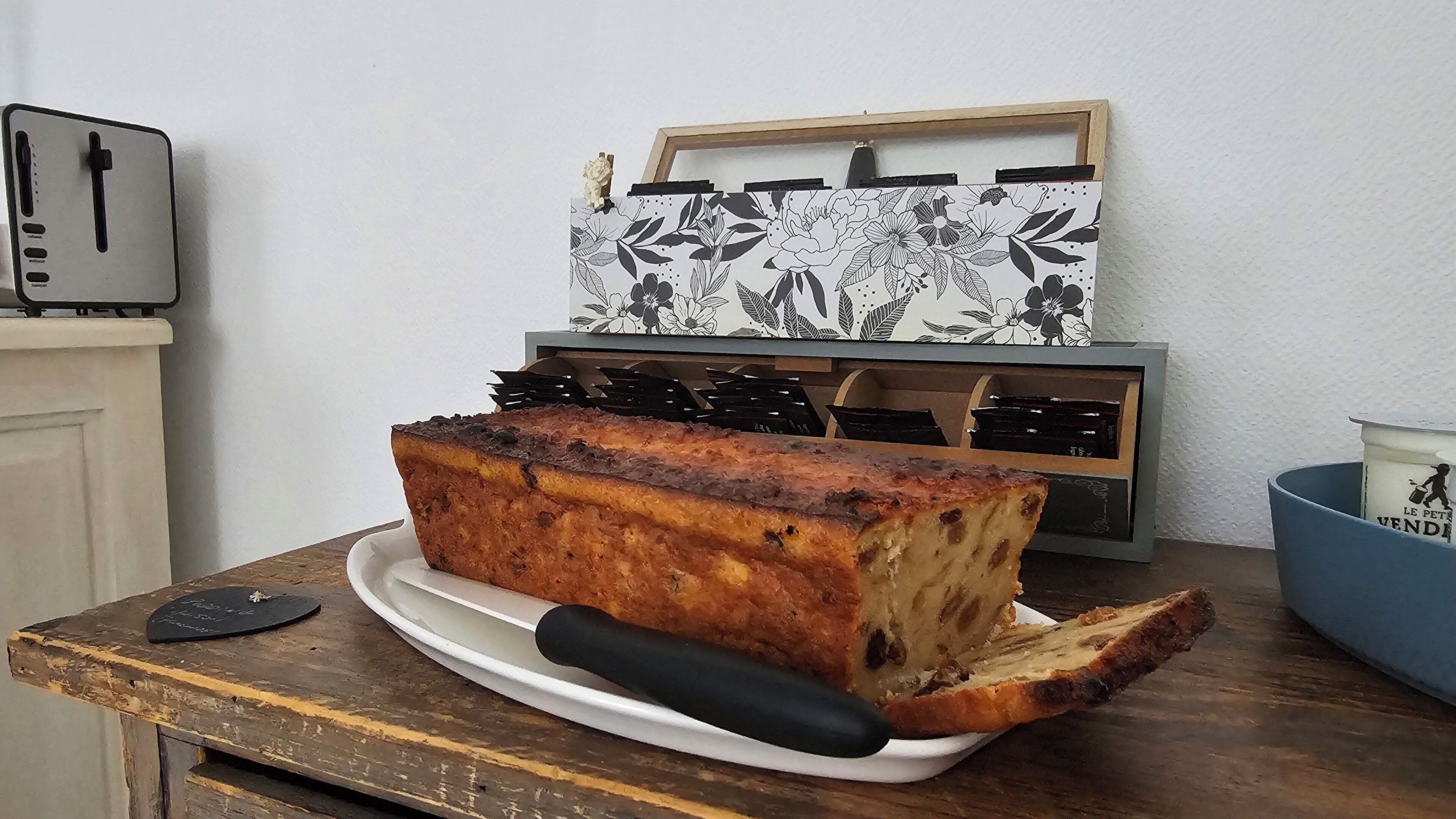 Una hogaza de pan sobre un plato blanco con un asa negra, una tostadora y un objeto decorativo con motivos florales.
