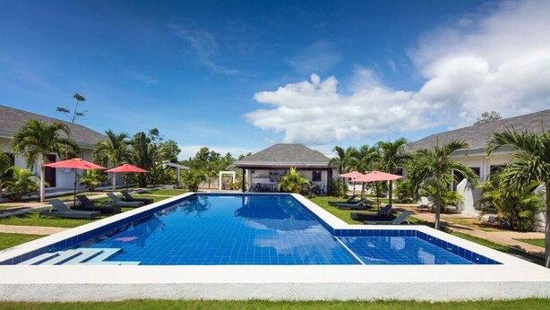 A swimming pool with lounge chairs and umbrellas, surrounded by palm trees and a building with a sloped roof. - ohana panglao resort (Panglao)