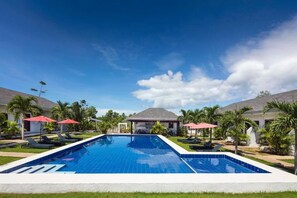 A swimming pool with lounge chairs and umbrellas, surrounded by palm trees and a building with a sloped roof. - ohana panglao resort (Panglao)