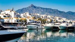 Un puerto deportivo con barcos amarrados, montañas al fondo y edificios blancos con tejados anaranjados.