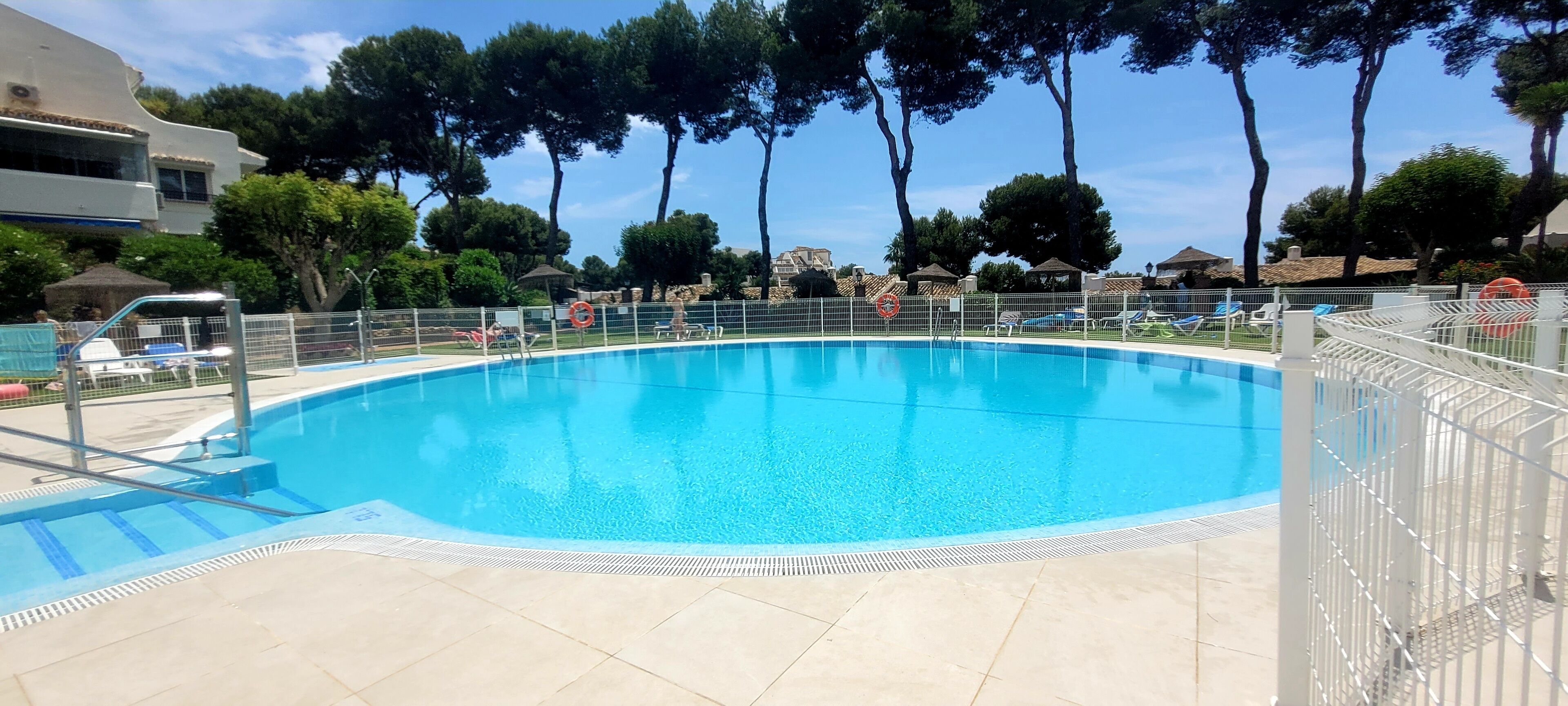 A swimming pool with a tiled deck, surrounded by a white fence, trees, and buildings in the background.