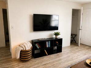 Una sala de estar con televisor de pantalla plana, una estantería, una planta pequeña, una cesta de mimbre y suelo de madera.