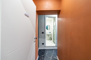 Ein schmaler Flur mit einer Tür zu einem Badezimmer. Das Badezimmer verfügt über einen Spiegel, ein Waschbecken und eine Toilette. Der Flur hat eine weiße Tür und orangefarbene Wände.