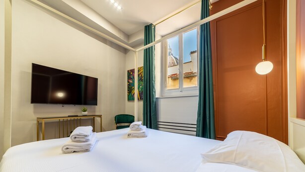 A hotel room with a bed, a flat-screen TV, a small table with a plant, and a window with curtains.