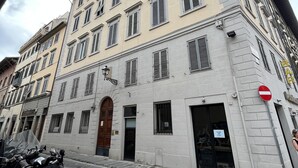 A multi-story building with a classic architectural style, featuring a central entrance with a wooden door, multiple windows with shutters, and a stone-paved street in front.